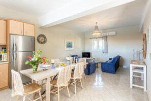 Una cocina moderna con refrigerador, una mesa de comedor y sillas. Hay una sala de estar con sofá y televisor.