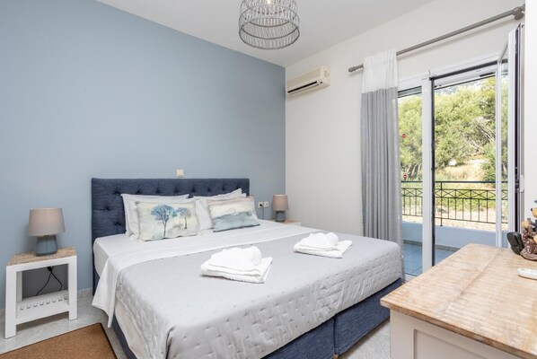 Un dormitorio con una cama grande, dos mesitas de noche, una lámpara de techo, un equipo de aire acondicionado, un balcón con vistas a zonas verdes y una ventana con cortinas.