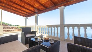 A covered outdoor seating area with wicker furniture and a table set for tea, overlooking a body of water. - House Green Side Sunset (Blato)