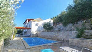 A stone-walled swimming pool with lounge chairs, a stone pathway, and a white building with a red roof. - House Green Side Sunset (Blato)