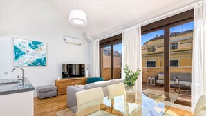 A modern living room with a glass dining table, grey sofas, a TV, and a large window with curtains.