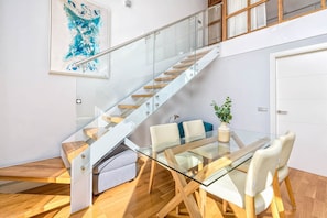 Un intĂ©rieur moderne avec un escalier en verre, un coin repas dotĂ© dâune table et de chaises en verre, et une porte blanche.