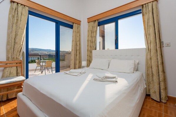 A hotel room with a large bed, two windows with blue frames, and a view of the landscape.