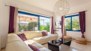 A modern living room with a white sofa, a glass coffee table, and a large window with purple curtains.