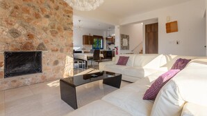 A modern living room with a stone wall, white sofas, a black coffee table, and a kitchen in the background.