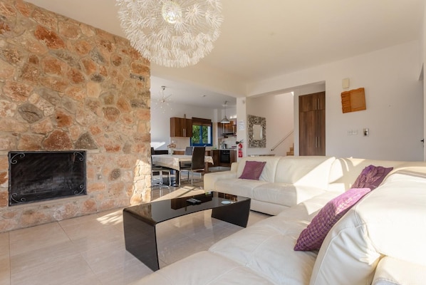 Un salón moderno con pared de piedra, sofás blancos, una mesa baja negra y una cocina al fondo.