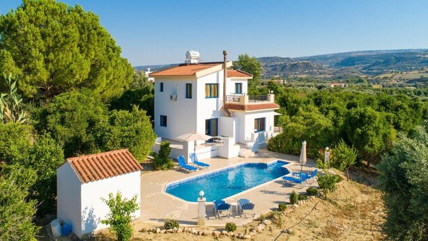 A two-story house with a swimming pool, surrounded by greenery and hills in the background.