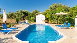 A rectangular swimming pool with a tiled deck, surrounded by blue lounge chairs and a white umbrella.