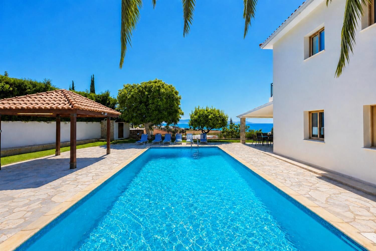En swimmingpool med solstolar och en pergola, omgiven av en vit byggnad och palmer.