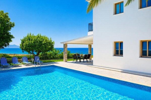 A swimming pool with lounge chairs, a covered patio, and a view of the sea.