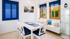A dining area with a white table, blue cushions, and white chairs. There are two windows with blue curtains, a painting on the wall, and a glass display case with items. - Argaka Sunset Villa - Three Bedroom Villa, Sleeps 6 (Argaka)