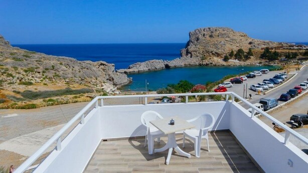 A balcony with a table and chairs overlooking a parking area and a rocky coastline.