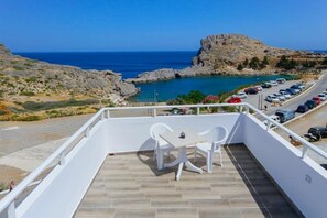 A balcony with a table and chairs overlooking a parking area and a rocky coastline. - St Pauls Bay Studios (Rhodes)