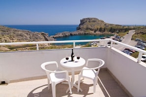 A balcony with a table and chairs, overlooking a coastal road and a rocky landscape with a view of the sea. - St Pauls Bay Studios (Rhodes)