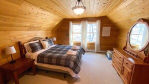 A cozy bedroom with a wooden ceiling, a bed with a plaid bedspread, a dresser with a mirror, and two windows with curtains.