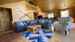 A cozy cabin interior with a wooden interior, a large sofa, a striped armchair, and a kitchen area.