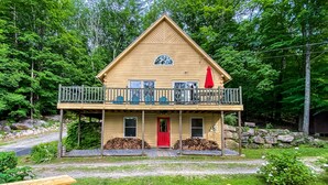 A two-story log cabin with a balcony, a red door, and a wooden deck.