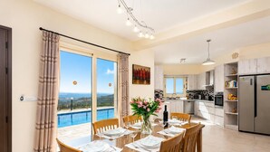 Comedor con mesa preparada para cuatro, cocina al fondo y vistas a una piscina y montañas a través de amplios ventanales.