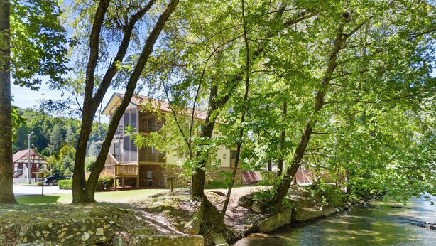 Un hotel a orillas del río con un puente de piedra, rodeado de exuberante vegetación y árboles.
