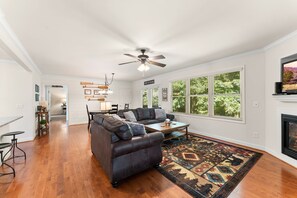 A spacious living room with a dark leather sectional sofa, a wooden coffee table, and a patterned rug. There is a ceiling fan, a fireplace, and large windows with a view of greenery.