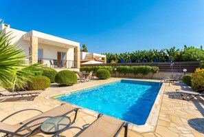 A rectangular swimming pool with lounge chairs and a patio area, surrounded by a stone wall and greenery.