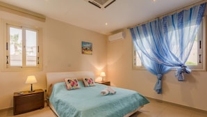 A bedroom with a bed, two bedside tables, a window with sheer curtains, and an air conditioning unit.