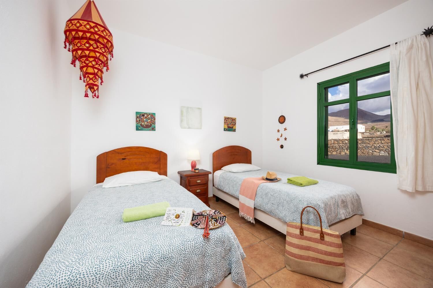Une chambre avec deux lits, une lampe suspendue, une fenêtre donnant sur un beau paysage et une petite table avec une lampe.
