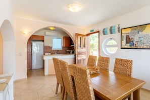 A dining area with a wooden table and woven chairs, a kitchen in the background, and a painting on the wall.