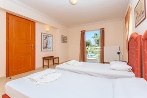 A hotel room with two beds, a wooden door, a small table, and a view of a pool and palm trees.