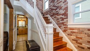 Una escalera con peldaños de madera y balaustres blancos, que conduce a un segundo piso con un muro de ladrillo y una ventana.