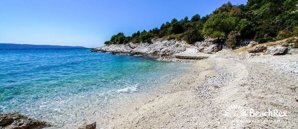 Enjoy a serene beach experience with clear waters and a pebbly shoreline, surrounded by a rocky cliff and lush vegetation.