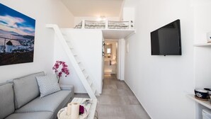 A modern, compact living space with a grey sofa, a staircase leading to a loft area, and a flat-screen TV mounted on the wall. - Christy Suites by Alpha Living (Mykonos)