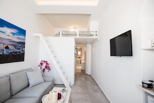 Un espacio habitable moderno y compacto con un sofá gris, una escalera que conduce a una zona superior tipo loft y un televisor de pantalla plana montado en la pared.