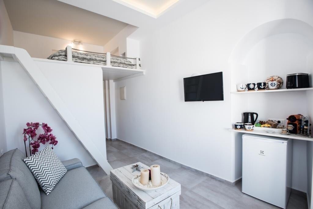 Een moderne, minimalistische woonruimte met een trap, een flatscreen televisie, een kleine keuken en een grijze bank met decoratieve kussens.