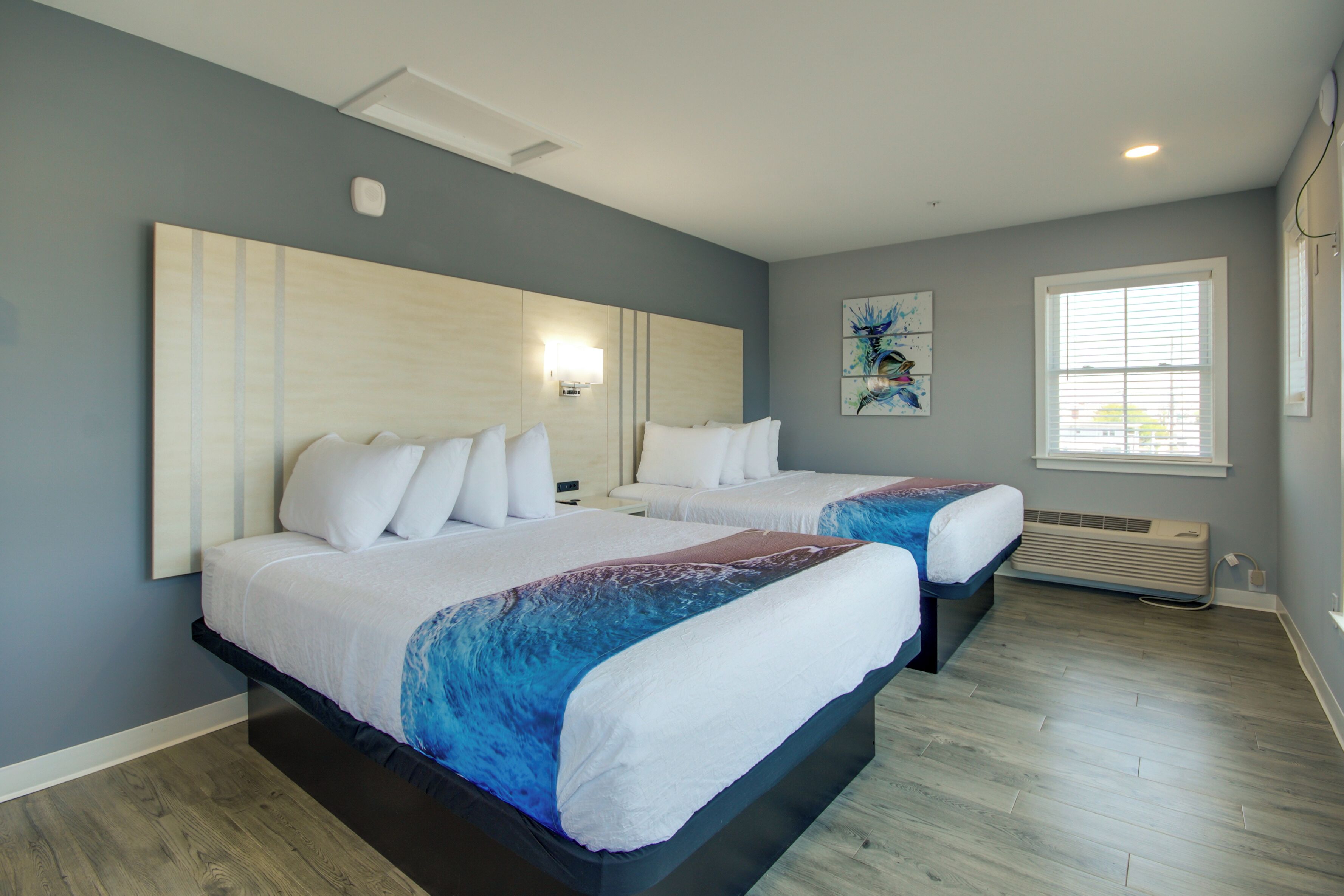 A hotel room with two beds, a large headboard, a wall-mounted light, a window with blinds, and a ceiling vent.