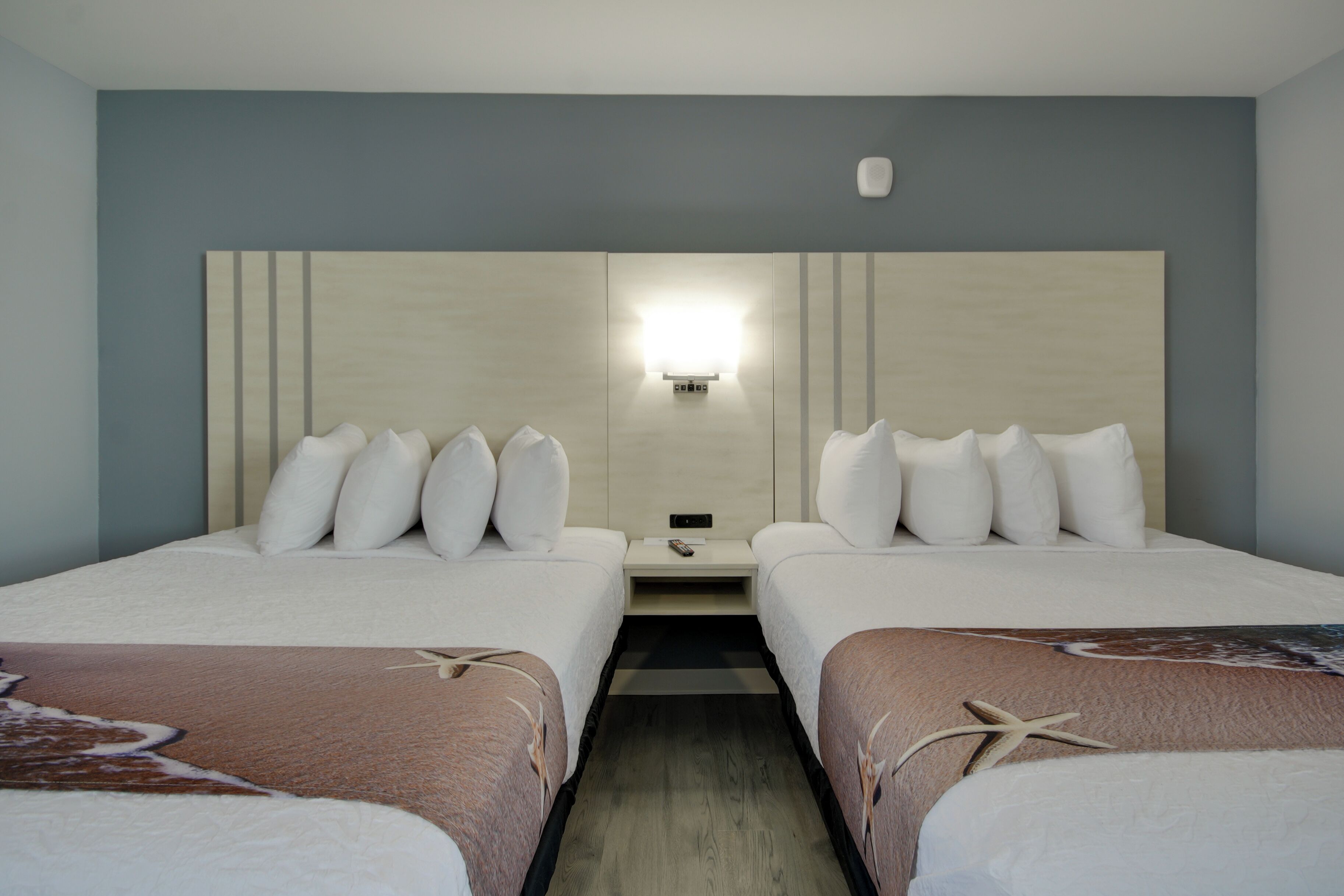A hotel room with two beds, a nightstand, and a wall-mounted light fixture.