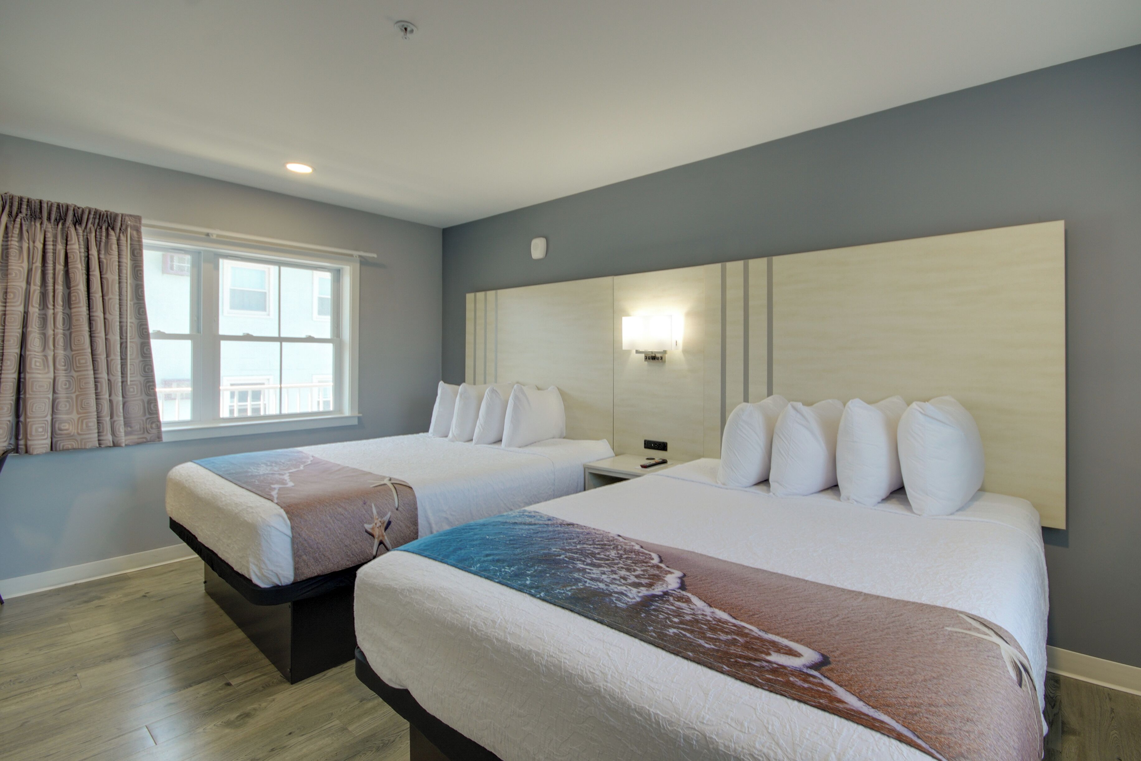 A hotel room with two beds, a large window with curtains, a wall-mounted light, and a wooden floor.