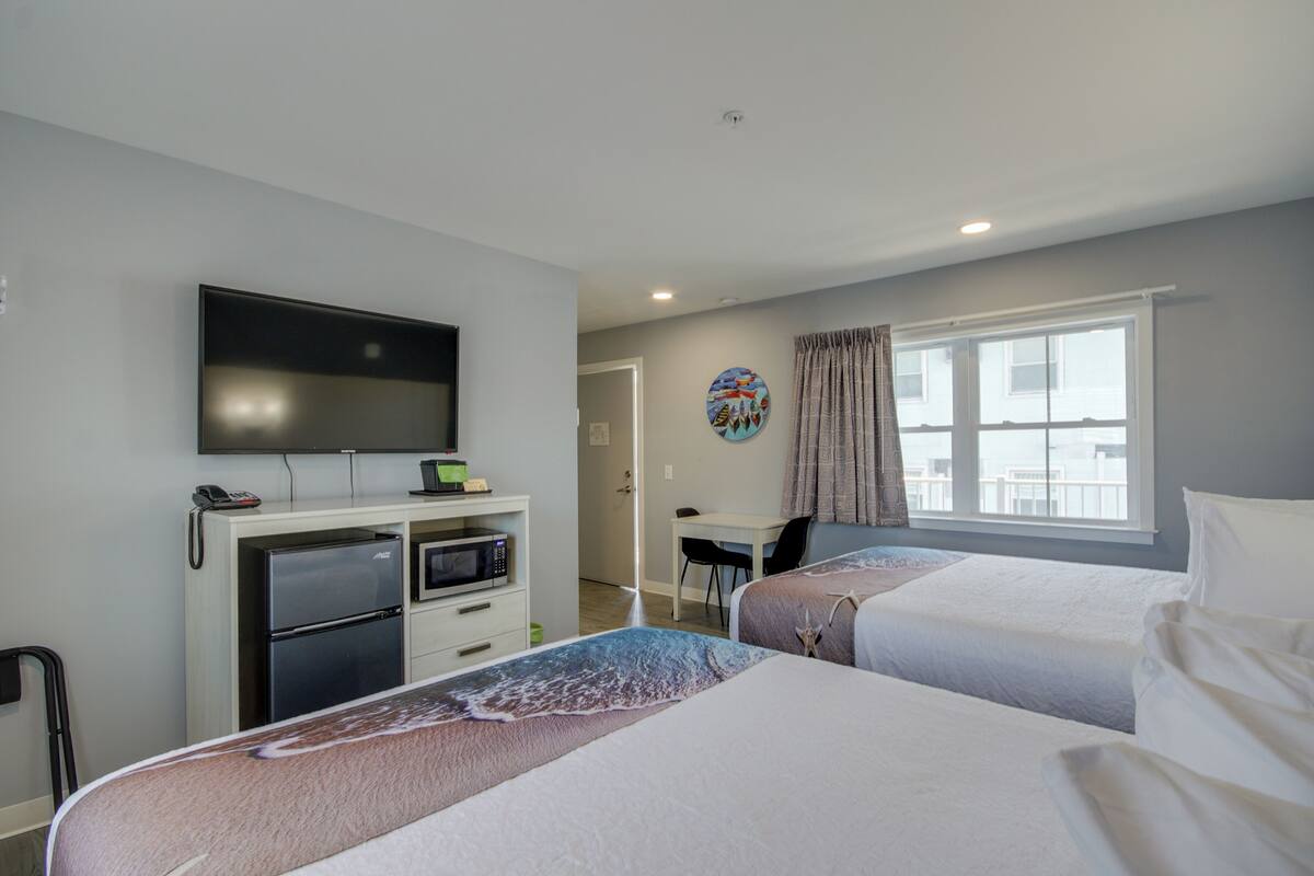 Ein Hotelzimmer mit zwei Betten, einem Fernseher, einem Kühlschrank, einer Mikrowelle, einem Schreibtisch und einem Fenster mit Vorhängen.