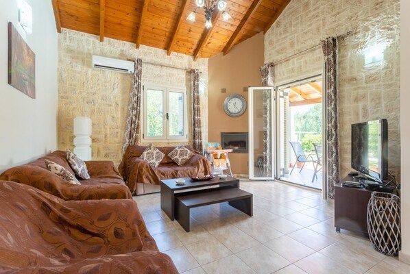 A cozy living room with a stone wall, wooden ceiling, and a large TV. - Villa Popi - Two Bedroom Villa, Sleeps 4 (Corfu)