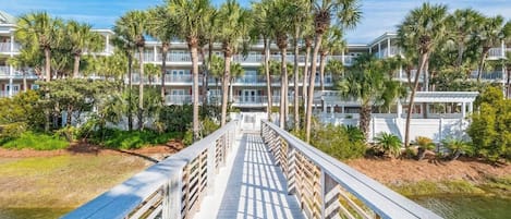 A boardwalk leading to a hotel with multiple floors, surrounded by palm trees and a body of water.