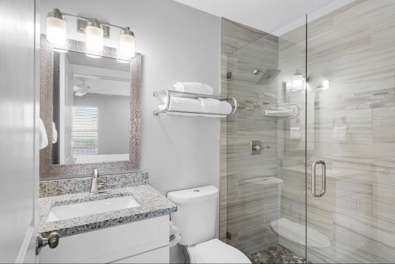 Une salle de bain moderne avec un lavabo surmonté d’un plan en granit, des toilettes blanches, une douche à parois de verre et un porte-serviettes.