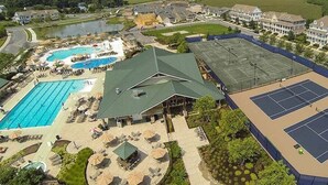 Vista aérea de un complejo turístico con piscina, canchas de tenis y un edificio con techo verde.