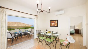 A dining area with a glass table and chairs, a chandelier, and a view of the outdoors. - Skala Villa Green - Three Bedroom Villa, Sleeps 6 (Ionian Islands)