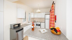 A modern kitchen with white cabinets, a microwave, a stove, and a refrigerator. There is a fruit bowl on the counter and a bottle of wine. - Skala Villa Green - Three Bedroom Villa, Sleeps 6 (Ionian Islands)