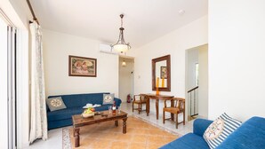 Ein Wohnzimmer mit einem blauen Sofa, einem Couchtisch mit Früchten und Gläsern, einem Esstisch mit Stühlen und einer Chandelüster.