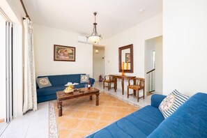 Una sala de estar con un sofá azul, una mesa de centro de madera con frutas y vasos, una mesa de comedor con sillas y una araña.
