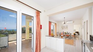 Eine moderne Küche mit einer zentralen Kochinsel, ein Essbereich mit Tisch und Stühlen sowie eine Glastür, die auf einen Balkon mit Meerblick führt.