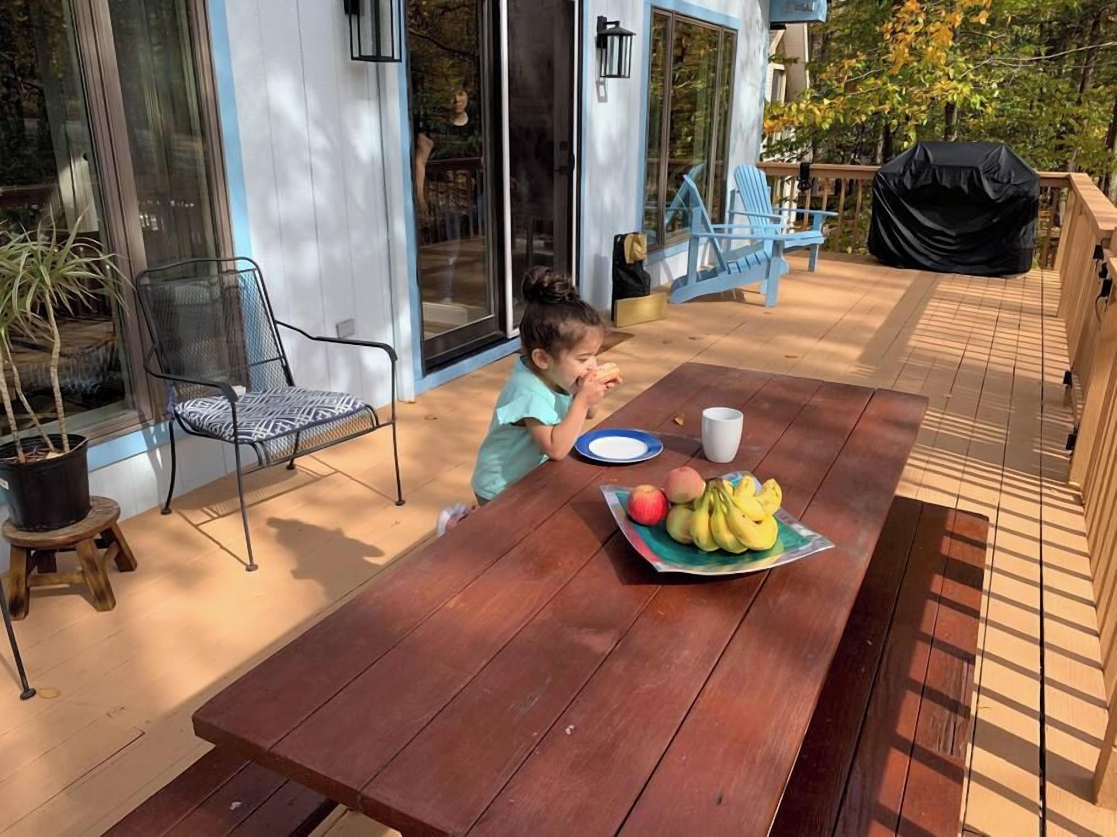 Een kind zit aan een houten buitentafel met een bord fruit en een beker, met een afgedekte barbecue en stoelen op de achtergrond.