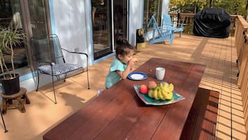 Een kind zit aan een houten buitentafel met een bord fruit en een beker, met een afgedekte barbecue en stoelen op de achtergrond.