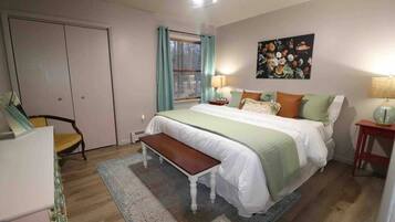 Un dormitorio con cama, banca, escritorio, silla, ventana con cortinas, un cuadro en la pared y ventilador de techo.